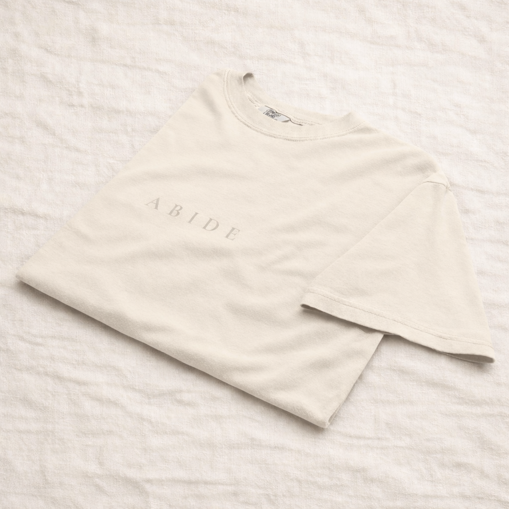 Cream folded unisex t-shirt with subtle wrinkles, featuring the word “ABIDE” printed in soft beige lettering, laid on a light textured fabric background.