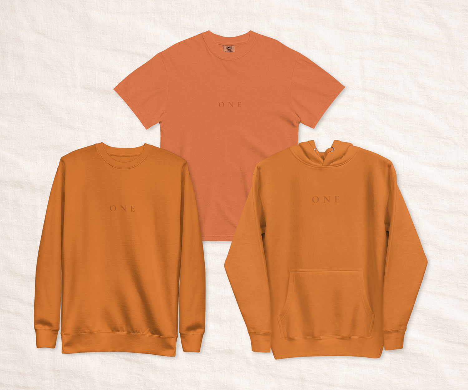 Three rust-toned unisex garments laid flat on a neutral fabric background, showing a T-shirt, crewneck sweatshirt, and hoodie with the ‘ONE’ wordmark to illustrate fit and colour across styles.