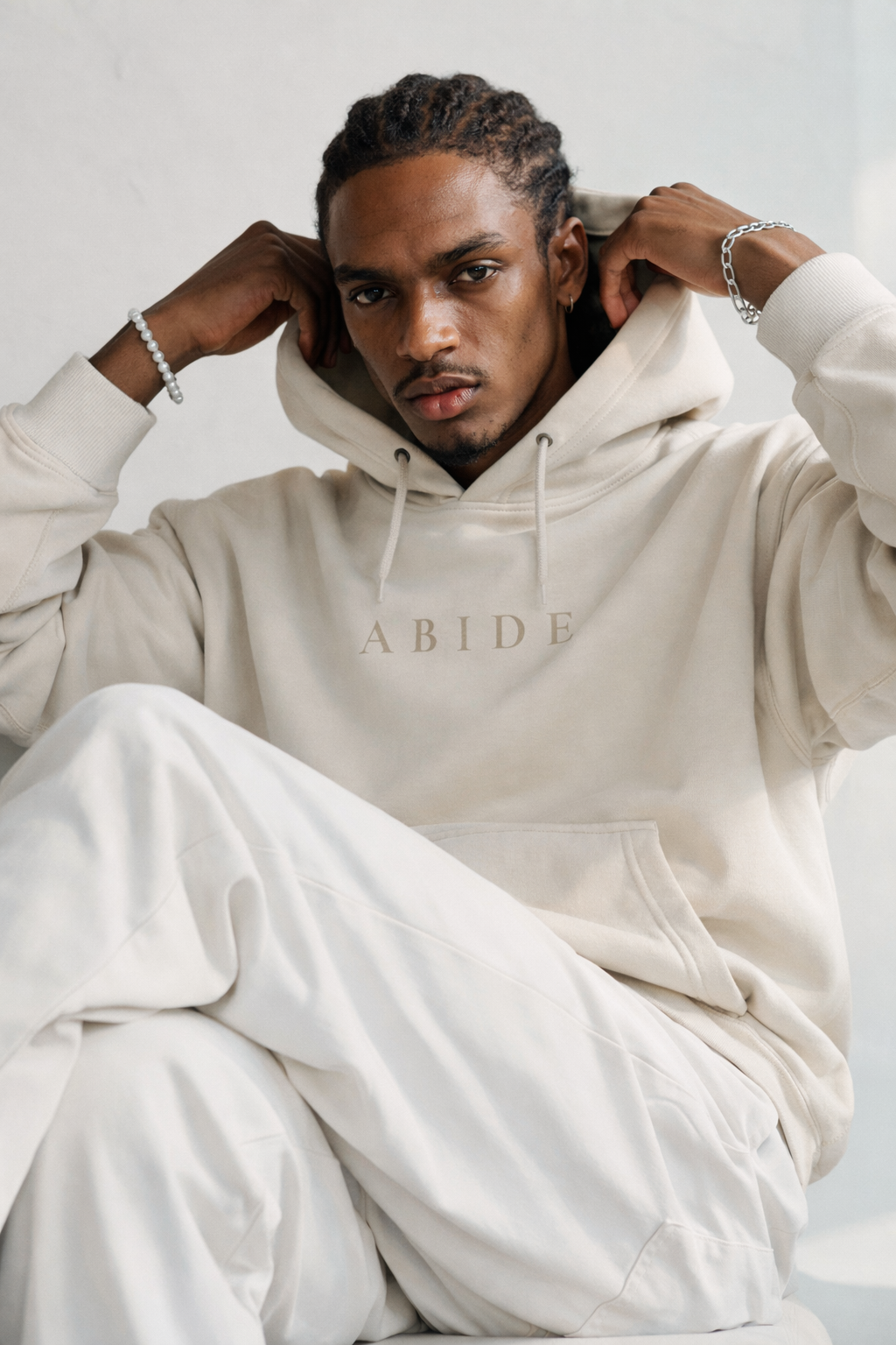 Model wearing a cream ABIDE hoodie with subtle tonal lettering, styled in a relaxed seated pose that highlights the soft drape and premium fabric of the garment.
